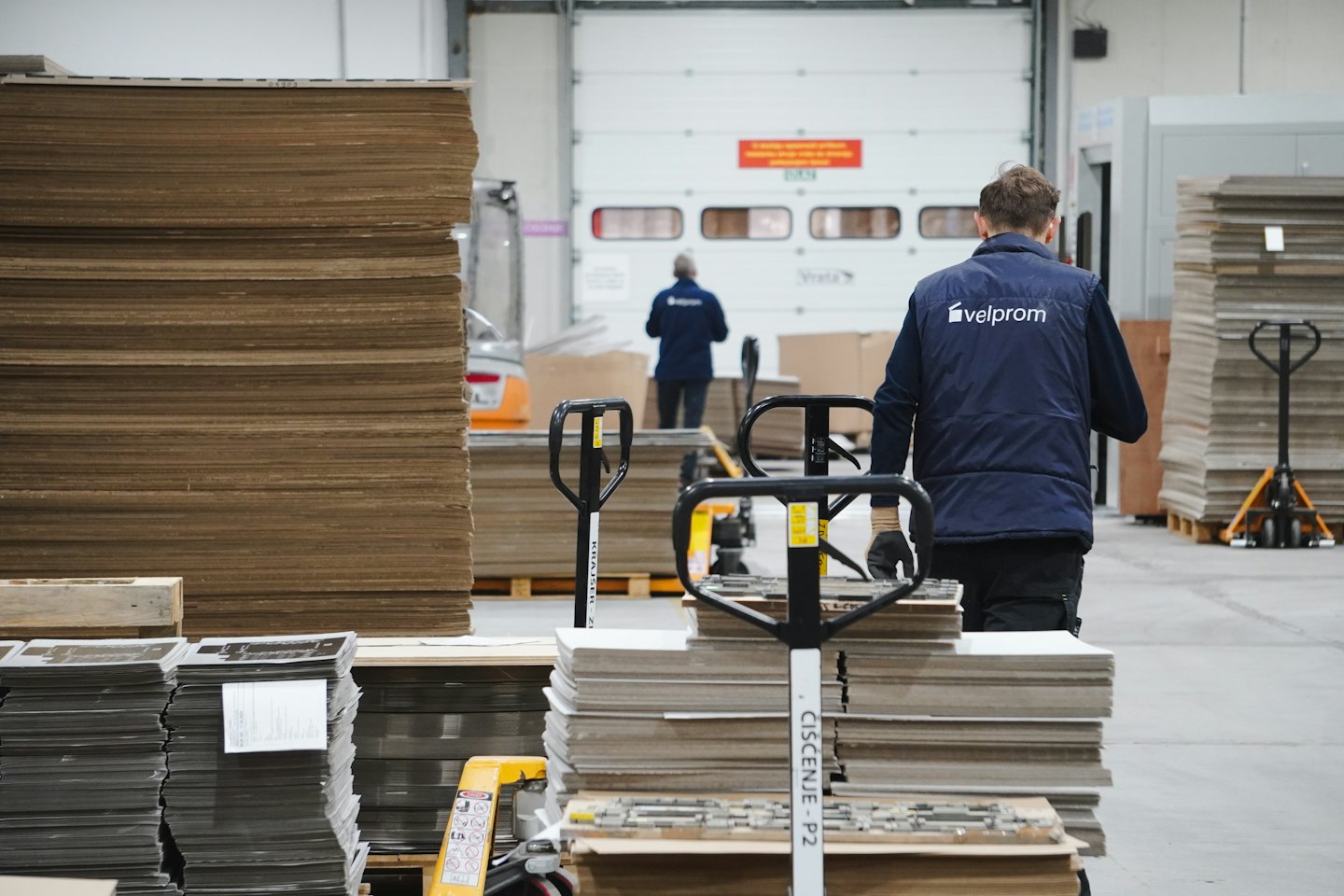 Hombres operan transpaletas en un almacén con cajas de cartón apiladas.